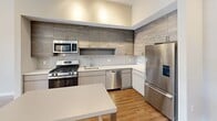 Located at Opus 410 in Stamford, CT, this two-bedroom, one-bathroom residence offers a well-designed layout with modern finishes throughout. The kitchen is a standout feature, equipped with stainless steel appliances including a gas range, microwave, dishwasher, and refrigerator. Textured gray cabinetry extends to the ceiling, complemented by a white tile backsplash, light countertops, and track lighting, creating a sleek and functional cooking space. A small dining table neatly bridges the kitchen and living areas.

Both bedrooms feature hardwood-style flooring and neutral-toned walls that provide a clean, versatile backdrop. The primary bedroom offers generous proportions and receives natural light, while the secondary bedroom includes sliding glass door access directly to the private outdoor patio — a defining feature of this unit.

The patio is a spacious, tiled outdoor area framed by stone-trimmed planters with lush greenery, offering a tranquil retreat with views of the surrounding landscaping and building architecture.

The bathroom is finished with a tub-shower combination, white tile surround, a stone-topped vanity, and a large LED-backlit mirror, delivering a clean and contemporary aesthetic throughout.