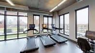 This fitness center combines an industrial-modern design with a comprehensive array of workout equipment, making it a fully functional facility for a wide range of training needs. The space is characterized by exposed concrete ceilings, visible ductwork, and structural concrete columns that lend it a raw, urban aesthetic, while linear LED lighting ensures the interior is well-lit throughout.

The main workout floor is generously proportioned, offering ample room for movement between equipment stations. Cardio equipment, including treadmills and stair climbers, is positioned along windowed walls, providing users with views of the surrounding cityscape. A dedicated strength training area houses a Smith machine, a full rack of barbells with weight plates, and a tiered dumbbell rack, catering to both beginners and experienced lifters.

A separate section of the facility is designated for functional training, featuring rowing machines, stability balls, and a wall-mounted television for guided workouts. An eye-catching abstract line-art mural adds a creative focal point to this area. A chalkboard wall and framed artwork near a small service counter contribute a welcoming, community-oriented feel.

Practical amenities include a dual water fountain and bottle-filling station set against a dark tile accent wall. Large black-framed windows and a glass wall opening to an outdoor terrace fill the space with natural light and provide a seamless connection to the exterior.
