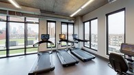 This fitness center combines an industrial-modern design with a comprehensive array of workout equipment, making it a fully functional facility for a wide range of training needs. The space is characterized by exposed concrete ceilings, visible ductwork, and structural concrete columns that lend it a raw, urban aesthetic, while linear LED lighting ensures the interior is well-lit throughout.

The main workout floor is generously proportioned, offering ample room for movement between equipment stations. Cardio equipment, including treadmills and stair climbers, is positioned along windowed walls, providing users with views of the surrounding cityscape. A dedicated strength training area houses a Smith machine, a full rack of barbells with weight plates, and a tiered dumbbell rack, catering to both beginners and experienced lifters.

A separate section of the facility is designated for functional training, featuring rowing machines, stability balls, and a wall-mounted television for guided workouts. An eye-catching abstract line-art mural adds a creative focal point to this area. A chalkboard wall and framed artwork near a small service counter contribute a welcoming, community-oriented feel.

Practical amenities include a dual water fountain and bottle-filling station set against a dark tile accent wall. Large black-framed windows and a glass wall opening to an outdoor terrace fill the space with natural light and provide a seamless connection to the exterior.