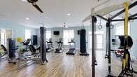 This fitness center offers a well-rounded exercise environment designed to support a variety of workout routines. The space features durable wood-look flooring throughout, light blue-toned walls, and recessed lighting complemented by ceiling fans that help maintain a comfortable atmosphere during workouts. Large windows along the perimeter invite ample natural light, creating an open and energizing feel.

The cardio section is equipped with multiple treadmills, elliptical trainers, and a stair climber, providing options for varied cardiovascular training. A wall-mounted flat-screen television is positioned for easy viewing during cardio sessions. Strength and functional training needs are addressed through a multi-station cable machine, a heavy punching bag suspended from the ceiling, and a functional training rig fitted with resistance cables and gymnastics rings. The combination of equipment supports everything from traditional strength work to boxing and bodyweight training. The overall layout is thoughtfully arranged to allow adequate spacing between equipment, promoting safe and efficient movement throughout the space.