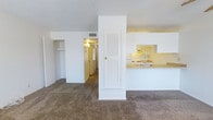 This single-floor residential space offers a functional and straightforward layout designed for comfortable everyday living. The living area features wall-to-wall carpeting in a neutral tone, creating a warm and cohesive feel throughout. A textured white ceiling adds subtle character to the space, while the bright white walls contribute to a clean, open atmosphere.

At the heart of the unit, a structural dividing column with a louvered closet door separates the living space from the kitchen area, providing a natural division without fully closing off the floor plan. The kitchen is positioned adjacent to the living area and features a pass-through countertop, light-colored cabinetry, and an under-cabinet range hood, making it both practical and accessible.

A hallway extending from the main living space leads to additional rooms, suggesting a well-organized layout that keeps private areas distinct from shared living spaces. Overhead ventilation is visible, indicating a centralized HVAC system. Built-in shelving is also visible, offering practical storage solutions integrated into the architecture. The overall ambiance of the space is bright, tidy, and move-in ready, with a neutral palette that provides a versatile backdrop for a variety of interior styles.