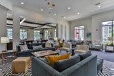 This single-floor amenity clubhouse is designed to impress, offering a thoughtfully curated collection of social and recreational spaces that blend contemporary style with welcoming comfort. The expansive game room is a standout feature, boasting multiple billiard tables, a well-appointed bar with counter seating and wall-mounted televisions, and large windows that flood the space with natural light while providing views of the surrounding community. Coffered ceilings with dark beam detailing add architectural character, complemented by mixed carpet and wood-look flooring throughout. The lounge area offers generous open-plan seating with an eclectic mix of mid-century modern furnishings, including bold yellow accent chairs, plush sectional sofas, and colorful artwork adorning the walls. Tall windows draw in abundant daylight and frame outdoor views, creating an airy and inviting atmosphere. A separate, more intimate sitting room features tufted velvet sofas, a bold graphic area rug, a linear fireplace set into a textured feature wall, and a wall-mounted television, making it ideal for relaxed gatherings. Pendant lighting, curated décor, and a cohesive modern aesthetic tie the entire space together into a dynamic and versatile community amenity.