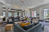 This single-floor amenity clubhouse is designed to impress, offering a thoughtfully curated collection of social and recreational spaces that blend contemporary style with welcoming comfort. The expansive game room is a standout feature, boasting multiple billiard tables, a well-appointed bar with counter seating and wall-mounted televisions, and large windows that flood the space with natural light while providing views of the surrounding community. Coffered ceilings with dark beam detailing add architectural character, complemented by mixed carpet and wood-look flooring throughout. The lounge area offers generous open-plan seating with an eclectic mix of mid-century modern furnishings, including bold yellow accent chairs, plush sectional sofas, and colorful artwork adorning the walls. Tall windows draw in abundant daylight and frame outdoor views, creating an airy and inviting atmosphere. A separate, more intimate sitting room features tufted velvet sofas, a bold graphic area rug, a linear fireplace set into a textured feature wall, and a wall-mounted television, making it ideal for relaxed gatherings. Pendant lighting, curated décor, and a cohesive modern aesthetic tie the entire space together into a dynamic and versatile community amenity.
