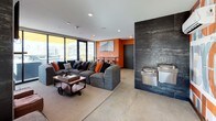 This single-floor communal lounge is designed with a bold, contemporary aesthetic that immediately draws attention. The space is anchored by vibrant orange accent walls that contrast sharply with large-format dark slate tile cladding featured prominently on a central architectural wall. This tile feature houses a stainless steel elevator and a built-in water fountain, adding both functional and visual interest to the layout.

Polished concrete floors run throughout, reinforcing the industrial character of the space. A generously sized grey sectional sofa serves as the social centerpiece, styled with orange and charcoal throw pillows that echo the room's color palette. An industrial-style coffee table on caster wheels adds a utilitarian charm to the seating area.

Opposite the seating arrangement, a dark tile fireplace surround frames a wall-mounted television, creating an inviting entertainment focal point. Floor-to-ceiling windows line one wall, flooding the space with natural light and offering expansive urban views, with direct access to an outdoor terrace equipped with a grill. A bold typographic mural adds graphic energy to the far wall. Bar-height seating and restroom access complete the well-appointed layout.