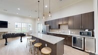 Located at 101 Cool Springs Dr in Elizabethtown, Kentucky, this single-floor amenity space is thoughtfully designed to serve as a central hub for socializing and relaxation. The kitchen area is a standout feature, showcasing rich dark-wood cabinetry, granite countertops with a textured geometric tile backsplash, and a large central island with bar stool seating illuminated by a trio of industrial-style pendant lights. Stainless steel appliances, including a French door refrigerator, microwave, and wine cooler, complete the well-equipped setup. Light hardwood-style flooring runs throughout, complementing the bright, neutral palette and contributing to the open, airy feel reinforced by soaring ceilings. A lounge area provides comfortable seating alongside a large wall-mounted television, a bar-height table with stools, and a foosball table, with large windows and French doors offering views of and access to an outdoor patio. A dedicated market nook features a stocked snack display and glass-door refrigerator for convenient 24-hour access. A connecting hallway ties the areas together, while curated abstract artwork adds a contemporary finishing touch throughout the space.