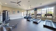 Located at 9303 E 81st St in Tulsa, Oklahoma, this fitness center offers a thoughtfully designed exercise space accommodating a comprehensive range of workout options. The room features durable rubber flooring throughout, providing a safe and functional surface for all types of exercise.

Large multi-pane windows line the walls, flooding the space with natural light and offering views of the surrounding outdoor greenery, creating an inviting and energizing atmosphere. Ceiling fans are positioned throughout, ensuring consistent airflow and comfort during workouts. Crown molding and wainscoting add a refined architectural touch that elevates the overall aesthetic above that of a typical gym.

The space is well-stocked with cardio equipment, including multiple treadmills and elliptical machines arranged along the windowed wall for an enjoyable workout experience. A dedicated free weights area features a tiered dumbbell rack with a full range of weights, accompanied by a weight bench and additional strength training equipment, including a multi-function cable machine. Wall-mounted televisions are strategically placed for easy viewing from various workout stations. Exercise reference charts posted on the walls provide useful guidance for users. A glass-paned interior door connects to an adjacent area, enhancing accessibility and flow within the facility.