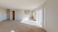 Located at 2445 Lyttonsville Rd in Silver Spring, Maryland, this single-floor unit offers a practical and well-organized layout. The open living area is a standout feature, filled with natural light streaming through large vertical-blind-covered sliding doors. Wall-to-wall carpeting adds warmth underfoot, while a ceiling fan provides year-round comfort. A pass-through window connects the living area to the kitchen, creating an open, communicative feel between the two spaces. The kitchen is equipped with warm wood cabinetry accented by sleek bar-pull hardware, stone-patterned countertops with generous L-shaped workspace, and a stainless steel sink. The bathroom features classic pink square tile work on the walls, maintaining a clean and timeless aesthetic. Adjacent to the bathroom is a spacious walk-in closet outfitted with a full-width hanging rod and shelf, offering ample storage and organization potential. The overall layout is efficient and functional, making practical use of every area within the single-floor footprint. Natural light plays a prominent role throughout, lending the interior a bright and airy atmosphere that enhances the neutral color palette found across all rooms.