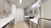 Located at 13525 Grayson Hill Circle in Dale City, VA, this well-designed one-bedroom, one-bathroom residence offers a thoughtful layout with modern finishes throughout. The kitchen is a standout feature, showcasing sleek gray shaker cabinetry with matte black hardware, crisp white quartz countertops, a subway tile backsplash, and a full suite of stainless steel appliances including a refrigerator, electric range, microwave, and dishwasher. A kitchen island with a pendant light and undermount sink adds both function and style. Light wood-look flooring runs through the kitchen and living areas, contributing to a clean, contemporary aesthetic. The living room is bright and airy, featuring neutral walls, a glass-paneled exterior door that invites natural light, and easy access to the outdoors. The bedroom is carpeted for comfort and includes a dedicated closet with built-in shelving and hanging rods, along with a window overlooking the surroundings. The bathroom features a full tub-shower combination with white tile surround and matte black fixtures, maintaining the modern design language found throughout the home. Overall, the space presents a cohesive, move-in-ready environment with a neutral palette and quality finishes.