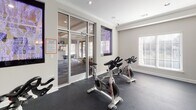 Located within the Integra Springs community at 4800 Integra Springs Boulevard in Kannapolis, NC, this dedicated Yoga/Spin Room is a purpose-built fitness studio designed to support a variety of workout routines. The single-floor space features durable dark rubber flooring, ideal for high-intensity cycling sessions and yoga practice alike, providing both grip and cushioning underfoot.

The room is outfitted with multiple Sunny spinning bikes arranged to accommodate group fitness sessions. A wall-mounted Panasonic flat-screen television provides on-demand workout content, making guided exercise sessions accessible directly within the room. A smaller wall-mounted display screen is also present for additional instructional use.

Crown molding and clean white walls give the studio a polished, well-maintained aesthetic. Natural light filters in through a large window fitted with white horizontal blinds, creating a bright and energizing atmosphere. Recessed lighting supplements the natural light, ensuring the space is well-lit at all times.

Floor-to-ceiling glass walls and a glass-paneled door open the studio visually to the adjacent community spaces, including a lounge and billiards area, fostering a sense of connectivity throughout the amenity floor. The overall design balances functionality with a refined finish, making it a standout fitness amenity within the Integra Springs community.