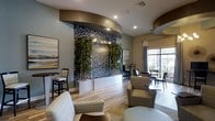 This single-floor business center is designed with a contemporary aesthetic that balances functionality and visual appeal. The space is immediately defined by a dramatic floor-to-ceiling water wall clad in mosaic tile, flanked by tall bamboo plants that introduce a natural, calming element to the environment. Warm-toned hardwood-style flooring runs throughout, tying together the various zones within the space.

Overhead, the ceiling features bold circular architectural accents finished in a rich bronze-toned trim, adding depth and dimension to the room. Recessed lighting and a decorative cluster chandelier provide layered illumination throughout the day and evening. Large windows along the perimeter invite abundant natural light and offer views to the outdoors.

The layout is thoughtfully divided into multiple functional areas. A comfortable lounge zone features upholstered striped armchairs and soft seating arranged for relaxed conversation or informal meetings. A dedicated workstation area is equipped with computer desks, ideal for productivity. Additional high-top bistro tables and standard dining-height seating provide flexible options for individual use or small group gatherings.

Decorative wall art and sunburst accents complement the modern design language throughout, creating a polished, welcoming atmosphere suited for both focused work and casual collaboration.