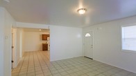 This single-floor space offers a clean, functional layout with a straightforward design. The main living area is finished with light-toned ceramic tile flooring that extends throughout, creating a cohesive and easy-to-maintain surface underfoot. Smooth white walls and a flush-mounted ceiling light fixture keep the space feeling bright and neutral. A white front entry door, accented by a semi-circular transom window above, allows a touch of natural light to filter into the entryway.

An open passage connects the living area to the kitchen, which is visible in the background. The kitchen features warm wood-toned cabinetry, a white range, and a white refrigerator, providing a functional cooking space with classic finishes. The recessed lighting in the kitchen adds subtle illumination to the work area. The overall layout promotes an open flow between the living and kitchen spaces, making the interior feel connected and accessible. The simple, uncluttered design of the space provides a versatile foundation that can accommodate a variety of furniture arrangements and personal styles.
