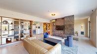 This elegantly designed single-floor amenity lounge offers a generous, open-plan layout that seamlessly blends living, lounging, and social spaces. The centerpiece of the main living area is a striking floor-to-ceiling feature wall clad in richly veined dark stone, which houses a mounted television and built-in shelving, creating a bold architectural focal point. Opposite, an expansive wall of floor-to-ceiling glass doors floods the interior with abundant natural light and opens directly onto a furnished outdoor terrace, offering views of the surrounding urban landscape. The lounge is appointed with a channeled tan leather sofa, a deep navy accent chair, and a substantial rectangular coffee table, all arranged over a textured area rug on polished light-toned tile flooring. A built-in bar and counter seating area, finished in warm wood tones with marble accents and brass hardware, provides an inviting space for gathering. A decorative open-grid shelving unit displaying curated art objects serves as a tasteful room divider, subtly separating the hallway and entry zone from the main lounge. Statement pendant lighting with gold interior detailing adds warmth and a refined finishing touch throughout, contributing to the space's overall sophisticated and contemporary ambiance.
