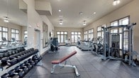 This fitness center is a comprehensively outfitted single-story facility designed to support a wide range of workout routines. The space is defined by its generous proportions, with high ceilings and an abundance of natural light streaming in through large, dark-trimmed multi-pane windows that line multiple walls. Views of the surrounding outdoor area, including what appears to be a pool courtyard, add a pleasant visual connection to the exterior environment.

The flooring throughout is durable, dark rubber tile — a practical and safety-conscious choice well-suited for heavy equipment and high-traffic use. The warm beige walls and recessed lighting contribute to a clean, functional ambiance.

Cardio equipment includes multiple treadmills and elliptical trainers arranged along the windowed walls, offering natural views during workouts. The strength training area is equally well-stocked, featuring a cable crossover machine, a functional trainer, a barbell rack with weight plates, adjustable benches, and an extensive dumbbell rack with a wide range of weights. Plate-loaded and selectorized strength machines round out the offerings.

A large wall mirror enhances spatial openness and provides visual feedback during training. A wall-mounted television adds entertainment convenience, making this a fully functional and thoughtfully arranged fitness facility.