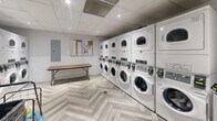 Located at 1465 Lawrence Ave W in Toronto, this shared laundry facility is a compact, efficiently designed space that accommodates multiple users with ease. The room is outfitted with a notable lineup of stacked front-load washers and dryers — each unit numbered for user convenience — providing ample laundry capacity. The interior features a striking herringbone-patterned light wood vinyl floor that adds a modern aesthetic touch, complemented by white subway tile wainscoting along the walls that contributes to a clean, bright atmosphere. A suspended drop ceiling with recessed pot lighting ensures the space is well-lit throughout. A folding table is positioned centrally, offering a practical surface for sorting and folding laundry. The room also includes a utility sink, a notice board for resident communications, and an electrical panel. A grey door with a glass panel provides access to and from the facility. The overall design strikes a balance between functionality and a refreshed, contemporary appearance, making it a well-appointed shared amenity for residents of the building.