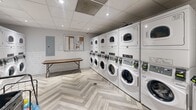 Located at 1465 Lawrence Ave W in Toronto, this shared laundry facility is a compact, efficiently designed space that accommodates multiple users with ease. The room is outfitted with a notable lineup of stacked front-load washers and dryers — each unit numbered for user convenience — providing ample laundry capacity. The interior features a striking herringbone-patterned light wood vinyl floor that adds a modern aesthetic touch, complemented by white subway tile wainscoting along the walls that contributes to a clean, bright atmosphere. A suspended drop ceiling with recessed pot lighting ensures the space is well-lit throughout. A folding table is positioned centrally, offering a practical surface for sorting and folding laundry. The room also includes a utility sink, a notice board for resident communications, and an electrical panel. A grey door with a glass panel provides access to and from the facility. The overall design strikes a balance between functionality and a refreshed, contemporary appearance, making it a well-appointed shared amenity for residents of the building.
