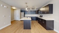 Located at 5397 W Borglum Ln in Herriman, Utah, this single-floor residence offers a clean, contemporary design throughout. The open-concept kitchen serves as the heart of the home, showcasing dark espresso cabinetry that contrasts elegantly with light quartz countertops. A central island with an undermount sink provides additional prep space and casual seating, while pendant lighting and a track light system illuminate the area with style. A full suite of stainless steel appliances — including a refrigerator, range, and microwave — completes the modern culinary setup. Light wood-tone flooring flows seamlessly from the kitchen into the adjoining living area, which benefits from a generous bank of windows fitted with horizontal blinds, allowing for ample natural light and a bright, airy atmosphere. The neutral warm-beige wall tones create a welcoming ambiance throughout. Two bathrooms are present, each featuring a tub-shower combination with chrome fixtures and tile-style flooring that adds a clean, polished finish. The layout is efficient and well-proportioned, with clearly defined zones for living, cooking, and personal retreat. Overall, this residence presents a well-balanced blend of modern finishes and functional design in a move-in-ready condition.