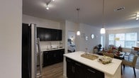 Located at 515 West Ave in Norwalk, Connecticut, this single-floor residence offers a well-designed open-concept layout with modern finishes. The kitchen is a standout feature, boasting dark cabinetry with sleek hardware, a white subway tile backsplash, and light quartz countertops that extend into a generous island. Stainless steel appliances, including a full-size refrigerator, complement the contemporary aesthetic. Pendant lighting hangs above the island, while track lighting illuminates the kitchen workspace, creating a well-lit and functional environment. The kitchen flows seamlessly into the living area, enhancing the open, airy feel of the space. Light wood-style flooring runs throughout, providing a warm contrast to the dark cabinetry. The neutral gray and white wall palette keeps the interior feeling bright and clean. A bedroom is visible from the main living area, offering a sense of connectivity within the floor plan. The overall ambiance is modern and polished, combining practical design with stylish details that make this a comfortable and inviting space.