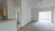 This single-story space is defined by its striking vaulted ceilings, which lend a sense of openness and volume to the interior. The soaring ceiling height is a standout architectural feature, creating an airy atmosphere throughout the main living area. A ceiling fan is mounted overhead, providing both function and a subtle design accent against the clean white ceiling.

The living area is finished with neutral-toned carpet flooring, contributing to a warm and comfortable feel underfoot. The walls are painted in a crisp, bright white, reflecting light effectively and enhancing the overall sense of space. A window positioned on the far wall allows natural light to filter into the room, adding to the bright and welcoming ambiance.

The layout is open and connected, with a partial wall or countertop visible to one side, suggesting an adjoining kitchen or breakfast bar area. A doorway visible in the background leads to additional rooms, one of which features louvered closet doors, likely a bedroom or utility area. The overall design is clean, minimal, and functional, offering a versatile canvas well-suited to a variety of furnishing styles.