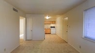 This single-floor space features a straightforward, functional layout designed for efficient living. The main living area is finished with wall-to-wall carpeting in a neutral beige tone, creating a soft and cohesive feel throughout. A textured ceiling adds subtle dimension to the otherwise clean, minimalist interior.

The kitchen is open and visible from the living area, separated by a partial wall that maintains a sense of connection between the two spaces. It is outfitted with light wood cabinetry providing ample storage above and below the counter. A white electric range and a sink with under-counter cabinetry complete the kitchen setup, while vinyl flooring offers a practical, easy-to-clean surface underfoot. Overhead lighting illuminates the kitchen area.

Multiple doorways are accessible from the main living space, leading to additional rooms or storage areas. The overall color palette is neutral throughout, with white walls and warm-toned finishes contributing to a bright and open atmosphere. The layout is practical and well-suited for everyday use, with each functional zone clearly defined within the compact footprint.