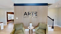 Located at 615 Tiny Town Rd in Clarksville, Tennessee, the Arte Luxury Townhomes clubhouse is a thoughtfully designed two-story amenity facility. The main floor welcomes residents through a set of glass double doors into a bright, open entry area with soaring ceilings, light hardwood-style flooring, and a branded accent wall featuring vertical wood slat paneling. The entry transitions seamlessly into a recreational lounge area anchored by a pool table, pendant lighting, and ample natural light from large windows. A staircase with a sleek metal railing leads to the upper level, while an open balcony maintains visual connectivity between floors. A dedicated living room offers a warm, modern retreat, complete with vertical wood slat accent walls, dark crown molding trim, a large wall-mounted television, a plush sectional sofa, and a ceiling fan. The adjacent gathering area features bold geometric-patterned wallcovering, a long communal table, and additional seating. The upper floor houses a well-equipped exercise room outfitted with rubber tile flooring, treadmills, ellipticals, stationary bikes, a stair climber, a weight bench, and a full rack of dumbbells alongside stability balls and resistance equipment — offering a comprehensive fitness experience for residents.