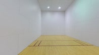 This space is a purpose-built indoor racquetball court designed to meet the demands of both recreational and competitive play. The court is defined by its smooth, light-toned hardwood flooring, finished to a polished sheen and clearly marked with regulation boundary lines and service zones in deep red, providing players with precise visual references during play. The enclosing walls are clad in large, seamlessly joined white panels that rise to a generous height, creating the expansive, unobstructed playing surface required for the sport. The tall ceiling contributes to an open, airy feel within the enclosed space. Overhead, recessed flush-mount light fixtures provide consistent, even illumination across the entire court, minimizing shadows and ensuring excellent visibility. At the rear wall, a horizontal observation window is positioned at an elevated level, allowing spectators to watch play from outside without interfering with the court environment. A utilitarian entry door is set into the back wall, providing access to the court. The overall design is clean, functional, and purpose-driven, with every architectural element oriented toward optimizing the racquetball playing experience.