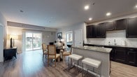 This single-floor residence offers a thoughtfully designed open-concept layout that seamlessly connects the kitchen, dining, and living areas. The kitchen is a standout feature, showcasing rich dark cabinetry, granite countertops, a classic white subway tile backsplash, and a full suite of stainless steel appliances. A peninsula with bar seating adds both functionality and style, flowing naturally into the dining space and living room beyond. The living room is bright and welcoming, with a sliding glass door that opens to a private balcony, allowing natural light to fill the space and providing pleasant outdoor views. Hardwood-style flooring runs throughout the main living areas, lending warmth and continuity to the open floor plan. The primary bedroom is generously proportioned, featuring plush carpeting, large windows, and a connected walk-in closet with built-in wire shelving, alongside a private bathroom finished with tile flooring. A second bedroom, also carpeted and naturally lit, offers flexible use as a guest room or home office. Neutral gray tones throughout the residence create a calm, cohesive aesthetic, while recessed lighting adds a clean, modern finish to each room.