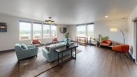 Located at 259 Ferncliff Ave NE on Bainbridge Island, WA, this single-floor communal space offers a thoughtfully designed open-concept layout that seamlessly blends comfort and modern aesthetics. The main lounge area is generously proportioned, featuring wide-plank hardwood flooring, smooth white ceilings with recessed lighting, and a striking multi-bulb statement chandelier. Multiple wall-mounted flat-screen televisions make this an ideal gathering space for both entertainment and socializing.

Large windows line the walls on multiple sides, flooding the interior with natural light while framing picturesque views of the surrounding Bainbridge Island landscape and outdoor terrace seating areas. The lounge is furnished with a mix of soft teal sofas, gray accent chairs with coral throw pillows, and functional side tables, creating an inviting and vibrant atmosphere.

Adjacent to the lounge, a kitchenette provides practical amenities including a full-size stainless steel refrigerator, a single-basin sink, and light wood cabinetry, all accented by a light blue subway tile backsplash. A large communal dining table surrounded by black chairs anchors the dining area, lit by two industrial-style cage pendant lights. Smaller bistro-style tables positioned near the windows offer additional seating with views of the surrounding neighborhood, making this space both functional and welcoming.