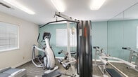 This fitness center at 5240 Rocklin Rd in Rocklin, California offers a well-organized, single-room workout space designed to accommodate a variety of exercise needs. The room features patterned carpet flooring and is finished with soft teal accent walls complemented by clean white walls, creating a fresh and energizing atmosphere. Flush-mounted ceiling lights provide bright, even illumination throughout the space.

The cardio section includes a treadmill and an elliptical trainer, positioned near windows fitted with horizontal blinds that allow natural light to filter in. The strength training area is anchored by a Life Fitness cable machine and a leg extension/curl bench, with a large wall mirror that visually expands the space and allows users to monitor their form during workouts.

Near the entrance, a wall-mounted flat-screen television provides entertainment during workouts, while posted fitness center rules and safety guidelines keep users informed. Sanitation stations with dispensers and waste bins are conveniently located by the entry, promoting cleanliness. The space opens into an adjacent area with hardwood-style flooring, framed artwork on the walls, and additional natural light, contributing to an inviting overall environment. Despite its compact footprint, this fitness center is thoughtfully equipped to support a well-rounded exercise routine.