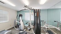 This fitness center at 5240 Rocklin Rd in Rocklin, California offers a well-organized, single-room workout space designed to accommodate a variety of exercise needs. The room features patterned carpet flooring and is finished with soft teal accent walls complemented by clean white walls, creating a fresh and energizing atmosphere. Flush-mounted ceiling lights provide bright, even illumination throughout the space.

The cardio section includes a treadmill and an elliptical trainer, positioned near windows fitted with horizontal blinds that allow natural light to filter in. The strength training area is anchored by a Life Fitness cable machine and a leg extension/curl bench, with a large wall mirror that visually expands the space and allows users to monitor their form during workouts.

Near the entrance, a wall-mounted flat-screen television provides entertainment during workouts, while posted fitness center rules and safety guidelines keep users informed. Sanitation stations with dispensers and waste bins are conveniently located by the entry, promoting cleanliness. The space opens into an adjacent area with hardwood-style flooring, framed artwork on the walls, and additional natural light, contributing to an inviting overall environment. Despite its compact footprint, this fitness center is thoughtfully equipped to support a well-rounded exercise routine.