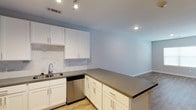 This single-floor space showcases a clean, modern design centered around an open-concept layout that connects the kitchen and living area. The kitchen is well-appointed with crisp white shaker-style cabinetry featuring gold-toned hardware, offering generous storage across both upper and lower cabinets. A neutral-toned countertop extends across the workspace and wraps around a peninsula, creating a natural division between the kitchen and the adjoining living space while also providing additional prep and seating space. The kitchen includes a stainless steel dishwasher, a single-basin sink with a contemporary faucet, and a tasteful tile backsplash that adds subtle texture to the space. Recessed lighting and a track light fixture illuminate the kitchen efficiently, maintaining a bright and airy atmosphere. Light hardwood-style flooring runs throughout the kitchen, transitioning into what appears to be a textured surface in the living area. The walls are painted in a soft, neutral blue-gray tone, complementing the white cabinetry and contributing to a calm, cohesive aesthetic throughout the space.