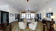 This well-appointed single-floor space combines comfort and functionality in a stylish communal setting. The open-plan layout is anchored by a central lounge area furnished with plush, cream-colored armchairs arranged around a small round coffee table, creating an inviting space for relaxed conversation or informal gatherings. A decorative, patterned carpet in warm earth tones adds visual interest and defines the central seating zone.

Elevated ceilings contribute to the sense of spaciousness, enhanced by a combination of recessed lighting and adjustable track lighting that runs along both sides of the room, allowing for versatile illumination throughout the day. A ceiling fan positioned centrally adds both style and air circulation.

Large double doors flanked by generously sized windows line the far wall, flooding the interior with natural light and offering views to the outdoors. Roman shades provide privacy and light control when needed.

Along the side walls, a series of dark-framed mirrors add depth and dimension to the space, while dining-style tables and chairs are neatly arranged, offering additional seating for meetings or events. The overall ambiance is clean, modern, and welcoming, making this space well-suited for a variety of social or professional uses.
