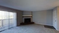 This single-floor space presents a functional and understated living area anchored by a corner fireplace as its central architectural feature. The fireplace is framed by a white mantel with a dark wood surround and raised hearth platform, adding a warm, traditional focal point to the room. The walls are finished in a neutral gray tone, creating a clean and versatile backdrop that complements a range of furnishing styles. Underfoot, wall-to-wall carpeting in a light gray-beige hue extends throughout, adding comfort and a cohesive, muted aesthetic to the space. The ceiling features a textured finish and is generously proportioned, contributing to an open, airy feel within the room. On the left side, a sliding glass door fitted with vertical blinds allows natural light to filter into the space while offering potential access to an outdoor area. The overall ambiance is quiet and neutral, making it a highly adaptable space suited to a variety of interior design preferences. The simplicity of the finishes and the open floor layout provide a solid foundation for personalization.