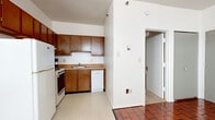This single-floor unit offers a straightforward and functional layout with a variety of distinct flooring materials that define each space. The most striking design element throughout the living and dining areas is the rich terracotta brick tile flooring, which adds warmth and character to the otherwise neutral white walls and textured ceilings. Multiple windows fitted with horizontal blinds allow natural light to fill these open areas, while baseboard heating runs along the perimeter walls to ensure year-round comfort.

The kitchen is efficiently designed, featuring warm oak cabinetry both above and below the countertops, providing ample storage. It is equipped with a stainless steel sink, a gas range with oven, a dishwasher, and a full-size refrigerator. The kitchen flooring transitions to a classic neutral vinyl tile, maintaining a clean and practical surface. An adjacent area continues the terracotta tile theme and benefits from large windows that overlook the surrounding neighborhood.

The bedroom offers a calm retreat with neutral carpeting underfoot and clean white walls, providing a comfortable and versatile space. The bathroom is compact and well-appointed, featuring light ceramic tile flooring, a wall-mounted sink, a toilet, and a bold black-framed mirror that adds a modern contrast to the all-white surroundings.