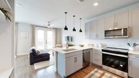 This thoughtfully designed studio offers an efficient, open-concept floor plan that seamlessly integrates the kitchen and living area into a cohesive, comfortable space. The kitchen is a standout feature, equipped with stainless steel appliances — including a refrigerator, range, microwave, and dishwasher — complemented by soft gray shaker-style cabinetry, a herringbone tile backsplash, and a clean white quartz peninsula with bar seating. A trio of matte black pendant lights hangs above, adding a modern touch. Rich hardwood-style flooring runs throughout, grounding the light, neutral color palette of the space. The living area benefits from abundant natural light streaming through floor-to-ceiling French doors, which also offer outdoor views and are dressed with elegant linen curtains. A ceiling fan adds both function and style overhead. The bathroom features a gray vanity with white countertop, a glass-enclosed shower, and coordinating hardwood-style flooring, delivering a clean and contemporary feel. A dedicated closet provides practical storage. Overall, the space presents a cohesive modern aesthetic with carefully selected finishes, making it a highly functional and visually appealing single-level studio.