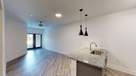 This single-floor unit offers a thoughtfully designed open-concept layout that seamlessly connects the kitchen and living area. The kitchen is equipped with stainless steel appliances, including a refrigerator, electric range, and over-the-range microwave, complemented by granite countertops, a subway tile backsplash, and sleek gray cabinetry with ample storage. A peninsula with a stainless steel sink and striking black pendant lights serves as both a functional workspace and a visual centerpiece. A built-in wine rack adds a refined touch to the kitchen design. The living area features light wood-style flooring that runs consistently throughout the unit, white walls, and a ceiling fan with a gold-toned fixture, lending a warm yet contemporary feel. Natural light filters in through a sliding glass door with views to the exterior. The bedroom is a comfortable, well-proportioned space with a window overlooking greenery, a ceiling fan, and direct access to a spacious walk-in closet outfitted with wire shelving on multiple levels. The bathroom is finished with a granite-top vanity, gray cabinetry, a framed mirror, and a tiled shower-tub combination. Paneled gray interior doors and clean white walls contribute to a cohesive, modern aesthetic throughout.
