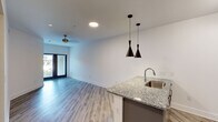 This single-floor unit offers a thoughtfully designed open-concept layout that seamlessly connects the kitchen and living area. The kitchen is equipped with stainless steel appliances, including a refrigerator, electric range, and over-the-range microwave, complemented by granite countertops, a subway tile backsplash, and sleek gray cabinetry with ample storage. A peninsula with a stainless steel sink and striking black pendant lights serves as both a functional workspace and a visual centerpiece. A built-in wine rack adds a refined touch to the kitchen design. The living area features light wood-style flooring that runs consistently throughout the unit, white walls, and a ceiling fan with a gold-toned fixture, lending a warm yet contemporary feel. Natural light filters in through a sliding glass door with views to the exterior. The bedroom is a comfortable, well-proportioned space with a window overlooking greenery, a ceiling fan, and direct access to a spacious walk-in closet outfitted with wire shelving on multiple levels. The bathroom is finished with a granite-top vanity, gray cabinetry, a framed mirror, and a tiled shower-tub combination. Paneled gray interior doors and clean white walls contribute to a cohesive, modern aesthetic throughout.
