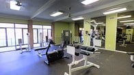 This single-floor fitness facility in Amarillo, Texas, offers a well-equipped workout space designed to support a range of exercise routines. The room features a textured ceiling with flush-mounted fluorescent lighting and a ceiling fan, ensuring a comfortable and well-lit environment. Large windows along one wall invite generous natural light and provide views of the surrounding outdoor area, creating an open and energizing atmosphere. The flooring consists of durable black rubber matting, ideal for a high-use fitness setting. Full-length mirrors line one wall, visually expanding the space and allowing users to monitor their form during workouts. The facility is outfitted with a variety of equipment, including a recumbent stationary bike, weight benches, a cable resistance machine, a leg raise station, and additional strength-training apparatus. Brick accent walls add a grounded, industrial character to the space, complementing the green-painted walls. The overall layout is open and functional, providing ample room to move between stations. This fitness space is practical and purposeful, offering a straightforward environment suited for both cardiovascular and strength-based training.