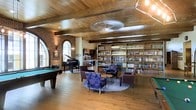 This single-floor space exudes a warm, sophisticated ambiance through a thoughtful blend of rustic and refined architectural details. The ceiling is a standout feature, clad in rich wood planking with exposed beams that lend a sense of craftsmanship and character throughout the room. A large, decorative ceiling medallion anchors the central space, adding a subtle industrial touch.

The expansive floor-to-ceiling bookshelf along one wall houses an impressive collection of books, establishing a strong library presence and serving as a natural focal point. Warm hardwood flooring runs throughout, complementing the wood ceiling and creating a cohesive, inviting atmosphere.

A grand piano is positioned near a striking arched window set into exposed brick, allowing natural light to filter in while adding historic architectural charm. Nearby, a billiards table provides a recreational counterpoint to the more contemplative library area. A comfortable seating arrangement—featuring armchairs and a patterned sofa—anchors a central lounge zone, making the space equally suited for relaxed conversation or quiet reading. Overall, the room balances function and elegance with notable warmth.