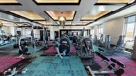 Elysian's amenity spaces are designed to deliver a resort-level experience. The main fitness center is a generously sized, single-floor facility distinguished by a coffered ceiling with warm ambient lighting and a striking chandelier centerpiece. The space is outfitted with a comprehensive array of strength and cardio machines — including cable systems, resistance machines, and free weights — arranged across vibrant, patterned modular carpet tiles in teal, pink, and charcoal. A dedicated cycling studio adjoins the main gym, featuring spin bikes, a wall-mounted display screen, built-in dark shelving, and a textured teal accent wall, with dark hardwood-style flooring adding a boutique-studio feel. The outdoor courtyard serves as the crown jewel of the amenity collection: a resort-style pool area anchored by a circular tufted lounge seat beneath a crystal chandelier and teal shade sails, accessible via a sweeping paved walkway framed by sculptural white arches, manicured greenery, and decorative art installations. A covered outdoor bar with a marble-topped counter and pergola overhead offers poolside views. A separate hammock lawn and branded hedge wall complete the al fresco experience, providing relaxed lounging areas within the landscaped grounds.