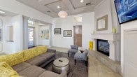 The Cross Pointe Clubhouse is a single-story community gathering space designed with a warm, contemporary aesthetic. The main lounge serves as the centerpiece, featuring a classic white-mantled fireplace, a large wall-mounted television, and a generous seating arrangement of gray sofas and patterned accent chairs positioned around a metallic drum coffee table. A coffered ceiling with a globe pendant light adds architectural interest and height, while recessed lighting and natural light from multiple windows enhance the open, welcoming atmosphere. Colorful artwork decorates the walls, complementing the space's yellow and neutral color palette.

Adjacent to the lounge, a compact kitchenette offers stainless steel appliances, white cabinetry with decorative panel detailing, a stone tile backsplash, and a white quartz bar counter with seating. Stylish black pendant lights hang above, adding a modern contrast to the bright space.

A separate café nook lined with floor-to-ceiling windows dressed in sheer curtains and wood blinds features bistro-height tables and sleek bar stools, bathed in abundant natural light. The combination of tile flooring throughout, bold yellow accent walls, and thoughtfully curated furnishings gives the clubhouse an inviting, community-focused ambiance.