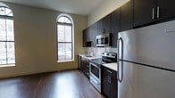 This single-floor space showcases a thoughtfully designed kitchen that serves as its centerpiece. The kitchen is outfitted with a full suite of stainless steel appliances, including a refrigerator, electric range, built-in microwave, and a undermount sink, all complemented by sleek, dark espresso cabinetry that extends to the ceiling for generous storage. White countertops provide a striking contrast against the dark cabinetry, while a white subway tile backsplash adds a clean, classic touch. The galley-style layout maximizes functionality and workflow efficiency. A tall, arched window with a distinctive divided-light design anchors one wall, drawing in abundant natural light and offering views of the surrounding streetscape. The warm-toned hardwood flooring runs throughout, grounding the space with richness and depth. The neutral wall color balances the contrast between the dark cabinetry and light countertops, creating a cohesive and modern aesthetic. The elevated ceiling height enhances the overall sense of openness, making the space feel airy and well-proportioned despite its focused layout.