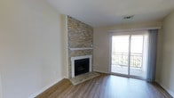 This single-floor space centers around a striking floor-to-ceiling stacked stone fireplace, which serves as the focal point of the main living area. The fireplace surround showcases warm, earthy tones of beige, gold, and grey stacked ledger stone, complemented by a clean white mantel and a tiled hearth that grounds the design. The combination of rustic stonework against the neutral cream-toned walls creates a balanced contrast between natural and contemporary elements.

The flooring throughout is a warm-toned, wood-style plank that adds continuity and warmth to the space. Large sliding glass doors fill the room with an abundance of natural light, opening onto a private balcony enclosed by a decorative metal railing, with views of surrounding greenery visible beyond. This seamless indoor-outdoor connection enhances the sense of openness and space within the living area.

The overall aesthetic is clean and understated, with neutral wall tones, white trim work, and recessed ceiling vents contributing to a polished, move-in-ready feel. The space is well-suited for comfortable everyday living, offering both a cozy focal point in the fireplace and an airy, light-filled atmosphere throughout.