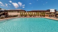 Located at 2418 Main St in Rocky Hill, Connecticut, this outdoor amenity space centers around a generously sized, crystal-clear swimming pool that serves as the focal point of the area. The pool features classic rectangular proportions with a clean tile finish and stainless steel entry rails, giving it a polished, resort-inspired aesthetic.

Surrounding the pool is an expansive concrete sundeck lined on all sides with rows of lounge chairs and shade umbrellas, providing ample space for relaxation and sunbathing. Lush landscaping, including neatly arranged columnar trees and ornamental plantings, frames the perimeter, enhancing privacy and adding a refined, manicured character to the setting.

A secure black metal fence encloses the pool area, with gated access points for safety. A water fountain station and safety life ring are conveniently positioned near the entrance. An elevated upper deck, accessible by staircase, overlooks the pool area and offers an additional outdoor gathering space with shaded seating.

The surrounding multi-story residential buildings provide a sense of community enclosure, while the open sky above allows for abundant natural light throughout the day. The overall ambiance is that of a well-appointed, thoughtfully designed outdoor retreat suited for warm-weather recreation and leisure.