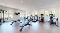 Located at 2708 Old Lebanon Pike in Nashville, TN, this fitness center offers a thoughtfully designed exercise space that accommodates a wide range of workout routines. The room is bathed in natural light, courtesy of large windows along multiple walls that provide open views of the surrounding greenery, creating an energizing atmosphere for exercise.

The space is outfitted with an impressive array of equipment. Cardio options include multiple treadmills, a rowing machine, a stationary bike, and an elliptical trainer. A multi-functional cable machine anchors one area of the room, ideal for strength and resistance training. A full rack of dumbbells is neatly organized alongside adjustable workout benches, catering to free weight exercises at various intensity levels. Wall-mounted medicine balls and a BOSU balance trainer round out the functional fitness offerings.

The interior is finished with clean white walls, recessed lighting, and durable flooring suited for high-activity use. Full-length mirrors are strategically placed to maximize the sense of space and support proper form during workouts. A wall-mounted television and a water station add convenience. The open, uncluttered layout ensures ease of movement between stations, making this fitness center a practical and well-appointed destination for residents pursuing their health and wellness goals.