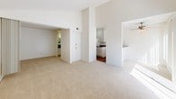 This single-floor space is characterized by its vaulted ceilings in the main living area, which lend an open, airy quality to the room. The living area is finished with neutral-toned carpeting and bathed in natural light from large sliding glass doors fitted with vertical blinds, creating a bright and welcoming atmosphere. A wall unit provides climate control, and a front entry door with a small hardwood threshold anchors the space.

The layout connects the living area to the kitchen, which features hardwood-style flooring, white cabinetry, and dark granite-style countertops. A white electric coil range with an overhead hood, along with ample upper and lower cabinet storage, provides a practical and clean cooking environment. A built-in white cabinet unit in the transition area offers additional storage.

The bathroom is well-appointed with a wide vanity topped with matching dark granite-style countertops, white cabinetry with brushed nickel hardware, and a large mirror with overhead lighting. A full-size tub and shower combo with a white surround, a toilet, and hardwood-style flooring complete the bathroom. The overall design of the space is clean, neutral, and functional.