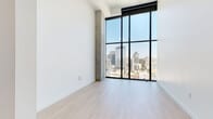 Located at 675 9th Ave in the heart of downtown San Diego, this contemporary high-rise unit delivers a refined urban living experience defined by clean lines, premium materials, and stunning city views. The open layout is anchored by dramatic floor-to-ceiling windows with bold black framing, flooding the interior with abundant natural light and offering panoramic views of the San Diego skyline. Light-toned hardwood flooring runs throughout, complementing the crisp white walls and exposed concrete structural columns that lend an industrial-modern character to the space. The chef's kitchen is a standout feature, equipped with high-end stainless steel appliances including a gas range, double wall oven, built-in microwave, wine refrigerator, and a full-size French door refrigerator. Soft-toned cabinetry extends to the ceiling for ample storage, while a white stone-topped island provides additional prep space and casual seating. A white subway tile backsplash adds a classic touch. The bathroom continues the sophisticated aesthetic with a floating wood-toned vanity topped in white stone, backlit mirror, warm brushed gold fixtures, and large-format tile flooring. In-unit laundry is also present. Every detail of this space reflects a commitment to contemporary design and elevated urban living.