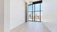 Located at 675 9th Ave in the heart of downtown San Diego, this contemporary high-rise unit delivers a refined urban living experience defined by clean lines, premium materials, and stunning city views. The open layout is anchored by dramatic floor-to-ceiling windows with bold black framing, flooding the interior with abundant natural light and offering panoramic views of the San Diego skyline. Light-toned hardwood flooring runs throughout, complementing the crisp white walls and exposed concrete structural columns that lend an industrial-modern character to the space. The chef's kitchen is a standout feature, equipped with high-end stainless steel appliances including a gas range, double wall oven, built-in microwave, wine refrigerator, and a full-size French door refrigerator. Soft-toned cabinetry extends to the ceiling for ample storage, while a white stone-topped island provides additional prep space and casual seating. A white subway tile backsplash adds a classic touch. The bathroom continues the sophisticated aesthetic with a floating wood-toned vanity topped in white stone, backlit mirror, warm brushed gold fixtures, and large-format tile flooring. In-unit laundry is also present. Every detail of this space reflects a commitment to contemporary design and elevated urban living.