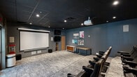 Located at 2508 SE Delaware St in Minneapolis, MN, this purpose-built screening room delivers a true theater experience in a compact, well-appointed space. The room is designed with a dark, cinema-inspired aesthetic, featuring deep blue-gray walls and a black drop-tile ceiling that enhance the immersive atmosphere. Recessed and track lighting provides controlled illumination throughout.

The seating area is arranged in tiered, stadium-style rows, with multiple rows of black leather theater chairs equipped with cup holders, ensuring comfortable viewing from every angle. A large retractable projection screen dominates the front wall, paired with a ceiling-mounted projector and a wall-mounted flat-screen display for additional viewing flexibility. A dedicated AV media cabinet below the screen houses supporting equipment, while wall-mounted surround sound speakers are positioned throughout for an enveloping audio experience.

A textured, patterned carpet covers the floor across all levels, adding warmth and acoustic dampening to the room. Near the entrance, a classic popcorn machine adds a fun, authentic movie-going touch. Double glass entry doors allow access from an adjacent space. A credenza with a wood-and-metal console table along the side wall adds a stylish, functional element. Decorative artwork displayed on the walls rounds out the thoughtfully designed interior.