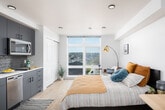 This thoughtfully designed studio unit offers a seamless open-plan layout that efficiently combines living, sleeping, and working areas within a single, well-organized space. Light wood-look flooring runs throughout, complementing the crisp white walls and clean-lined contemporary finishes.

The sleeping area is anchored by a full-sized bed and is paired with a dedicated workspace featuring a natural wood desk and ergonomic chair. A stylish sliding barn door provides a practical spatial divider, adding a modern industrial touch to the interior. One of the most notable features is the expansive floor-to-ceiling window that floods the room with natural light while framing sweeping views of the surrounding cityscape and neighborhood rooftops.

The kitchenette is fully equipped with stainless steel appliances, including a refrigerator, microwave, dishwasher, and cooktop. Blue-gray cabinetry with matte black hardware pairs with a textured tile backsplash to create a stylish yet functional cooking space. Ample upper and lower cabinet storage is provided throughout.

The bathroom includes a walk-in shower finished in clean white surround with matte black fixtures, maintaining a consistent and cohesive modern aesthetic. Recessed lighting throughout the unit ensures a bright, welcoming ambiance at any time of day.