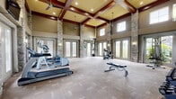 Located at 15180 Old Hickory Blvd in Nashville, TN, this fitness center is a standout facility defined by its dramatic architectural design and spacious, open layout. The room commands immediate attention with its soaring vaulted ceilings adorned with rich, dark wood coffered beams and recessed lighting, creating a sense of grandeur rarely found in a fitness setting. Stately stacked-stone columns anchor the four corners of the space, adding a refined, lodge-inspired aesthetic that pairs naturally with the warm neutral tones of the walls and carpeted flooring. Ceiling fans suspended from the beams provide additional airflow throughout the room. An abundance of natural light pours in through multiple sets of glass French doors and high clerestory windows, offering views of the outdoor pool area and surrounding grounds. The fitness center is equipped with a comprehensive selection of exercise equipment, including treadmills, ellipticals, a recumbent bike, a cable machine, a weight bench, and a dumbbell rack, accommodating both cardio and strength training needs. Wall-mounted mirrors expand the sense of space and assist in form monitoring. A mounted television adds a modern convenience for entertainment during workouts. The overall ambiance is upscale yet functional, blending architectural elegance with a fully equipped fitness environment.